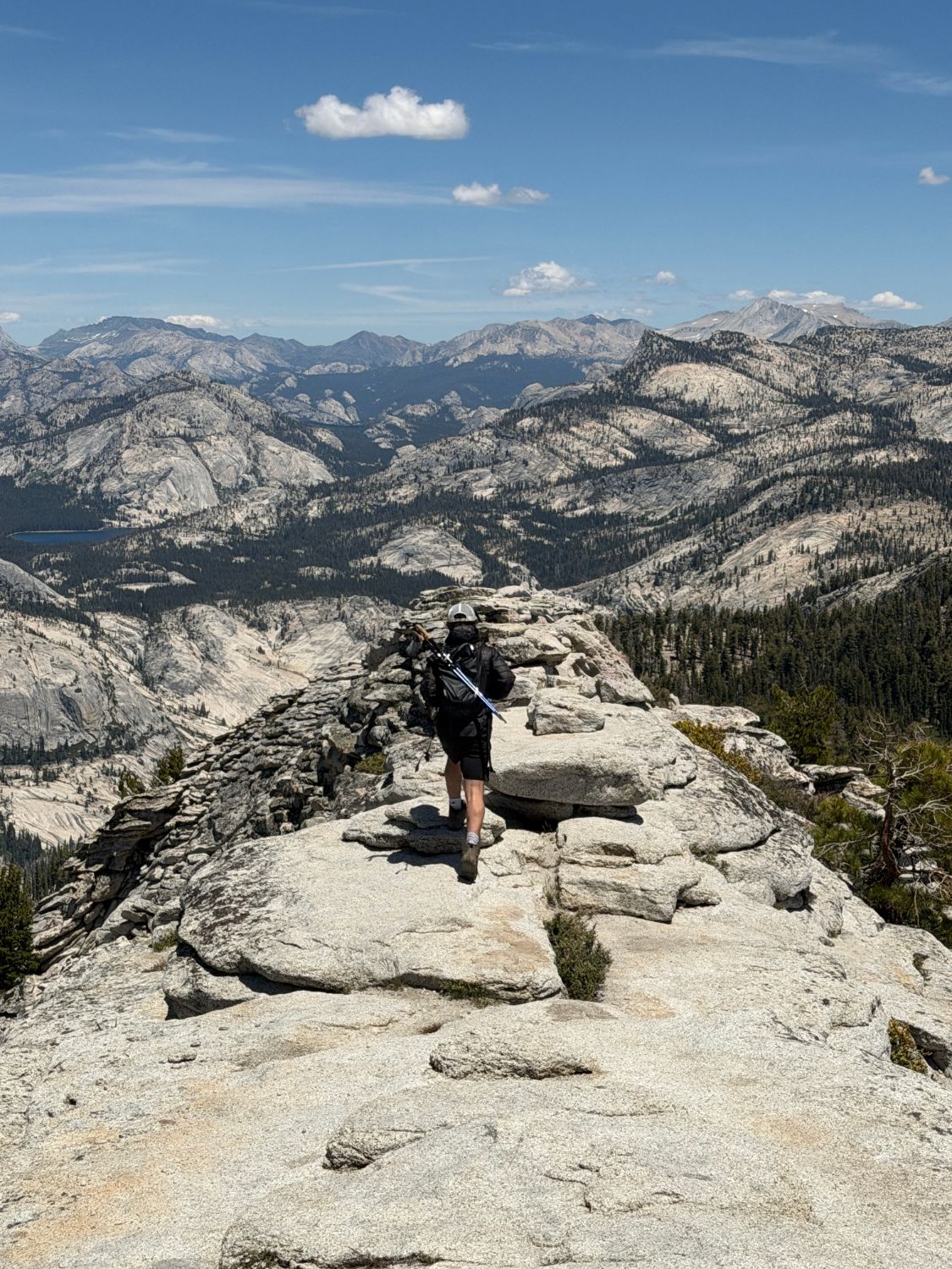 A hiker with trekking poles walking a granite ridge toward a vast Sierra Nevada landscape and a distant alpine lake