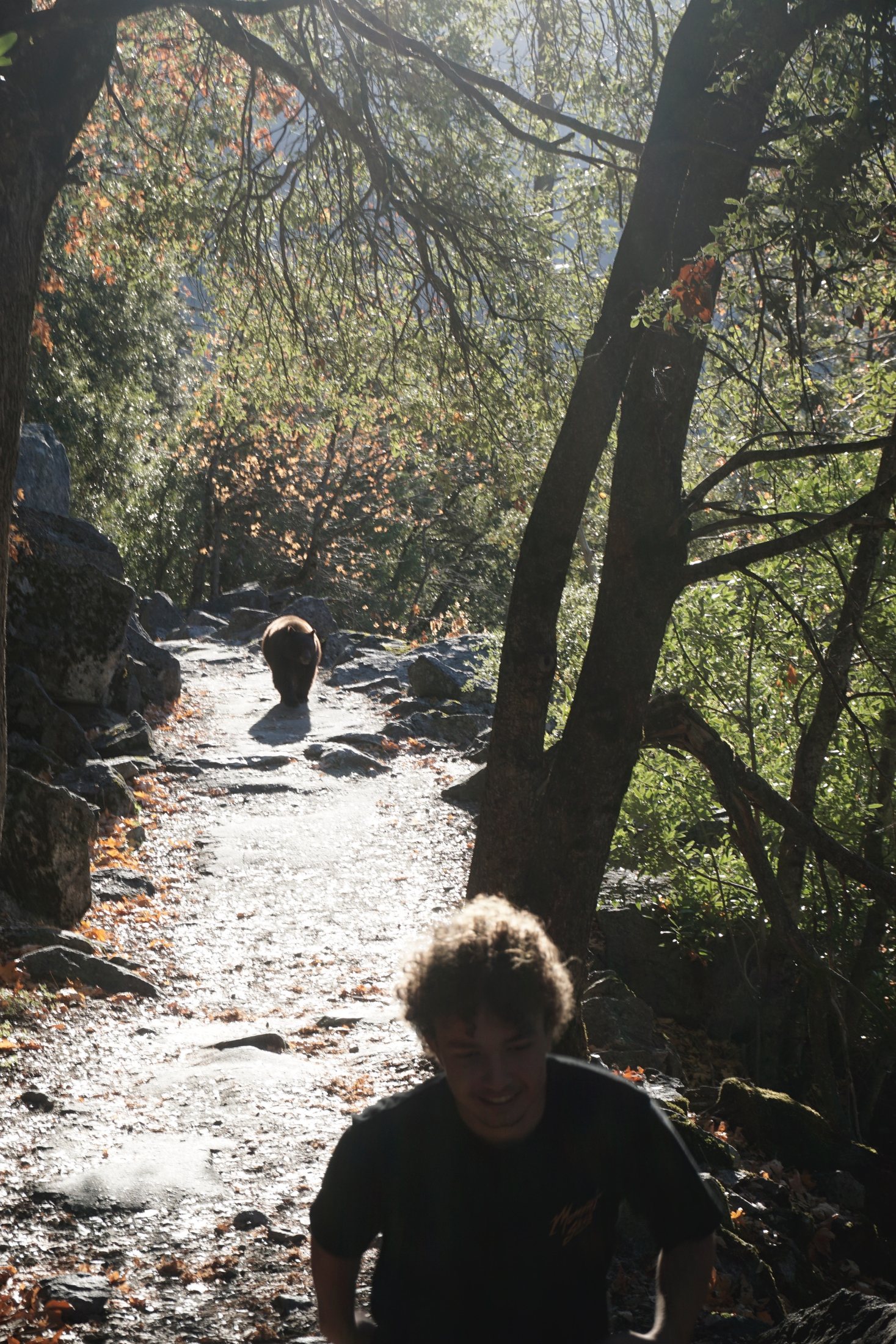 Marc grinning on a sunlit granite trail in autumn while a black bear walks along the path behind him