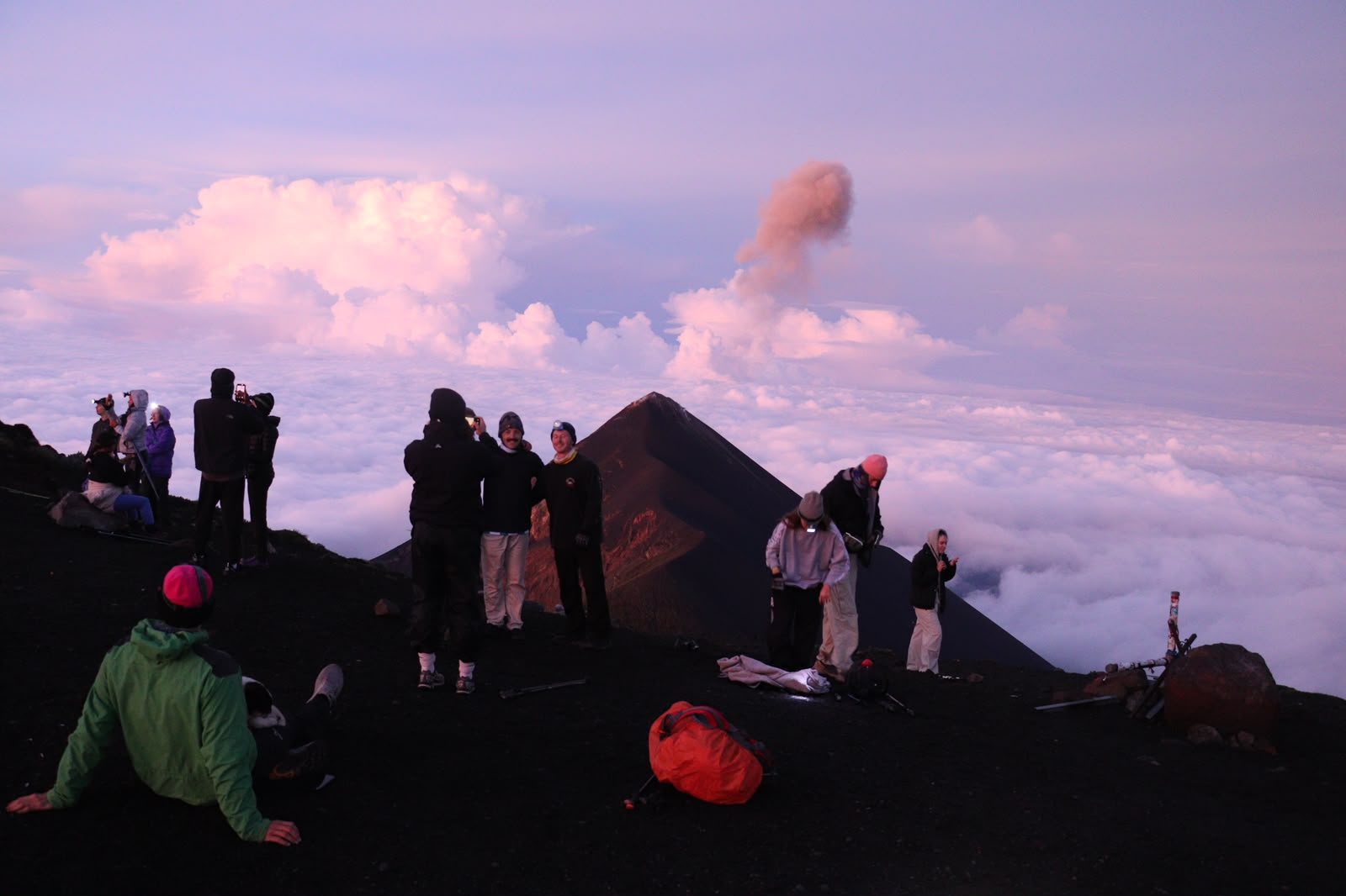 Hikers on a dark volcanic ridge at dawn watching Volcán de Fuego release a plume of ash above a sea of pink clouds