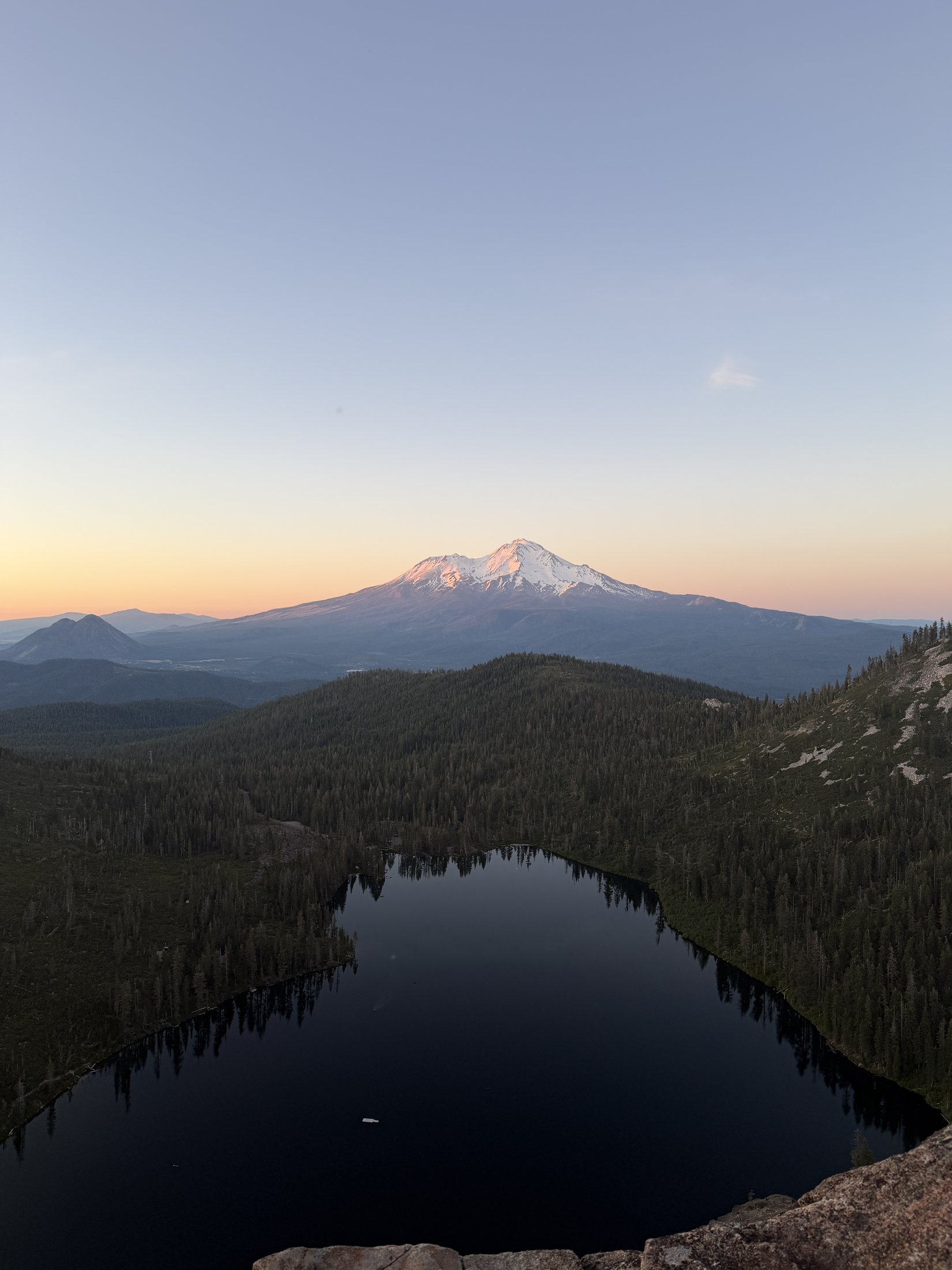 Snow-capped Mount Shasta glowing at golden hour above a forested ridge and a dark mountain lake