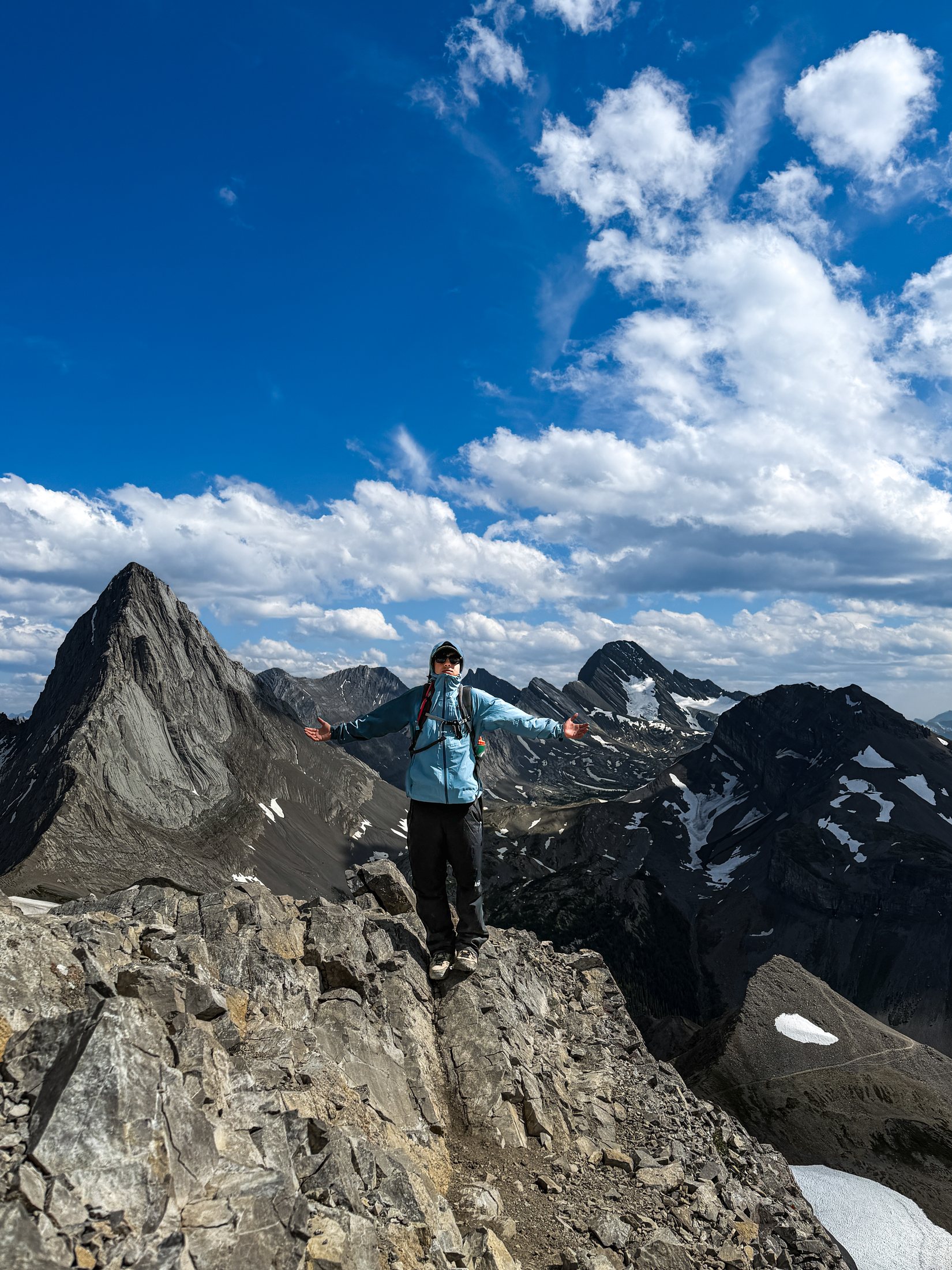 Marc standing on a rocky summit ridge with arms outstretched, a sharp limestone peak and snowfields behind him